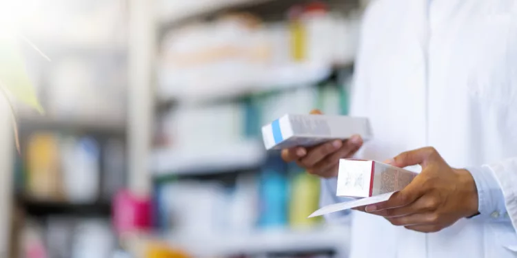 Closeup hand of woman pharmacist with prescription and medicine at drugstore. Photo of Woman pharmacist holding prescription checking medicine in pharmacy - drugstore. Single pharmacy technician in white lab jacket holding prescription scrip and generic medication box with shelf in background.