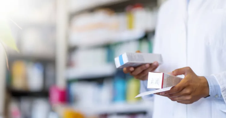 Closeup hand of woman pharmacist with prescription and medicine at drugstore. Photo of Woman pharmacist holding prescription checking medicine in pharmacy - drugstore. Single pharmacy technician in white lab jacket holding prescription scrip and generic medication box with shelf in background.