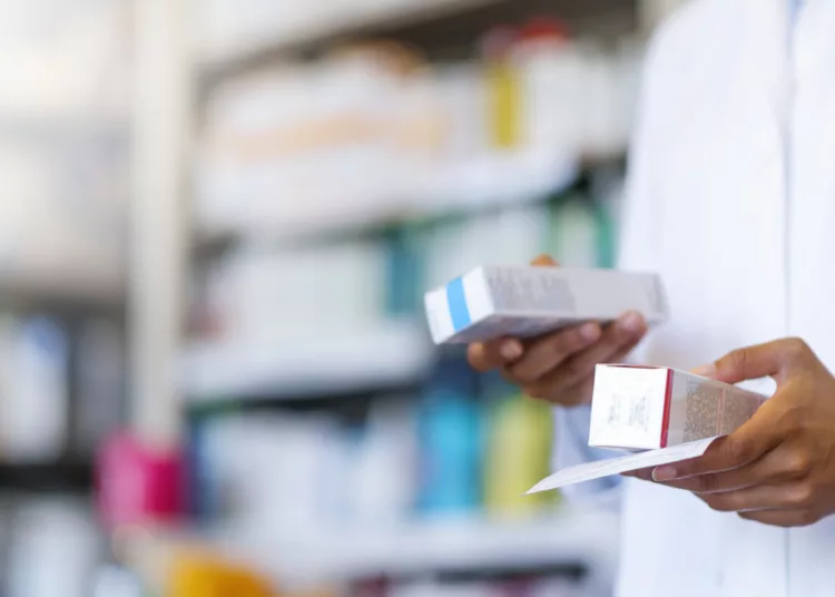 Closeup hand of woman pharmacist with prescription and medicine at drugstore. Photo of Woman pharmacist holding prescription checking medicine in pharmacy - drugstore. Single pharmacy technician in white lab jacket holding prescription scrip and generic medication box with shelf in background.