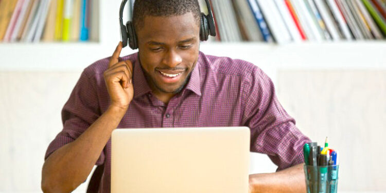 Smiling african american office worker in headphones looking at laptop screen. Young  casual businessman studying foreign language, communicating with clients through video conference application.