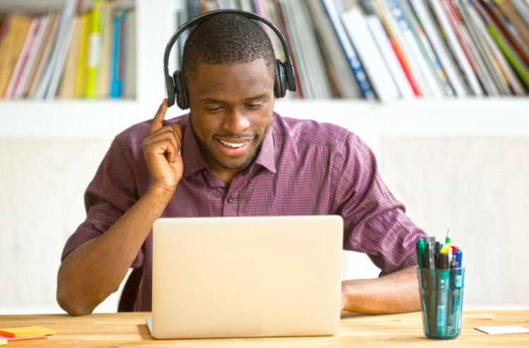 Smiling african american office worker in headphones looking at laptop screen. Young  casual businessman studying foreign language, communicating with clients through video conference application.