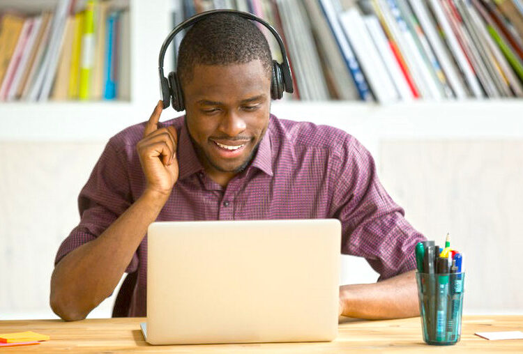 Smiling african american office worker in headphones looking at laptop screen. Young  casual businessman studying foreign language, communicating with clients through video conference application.