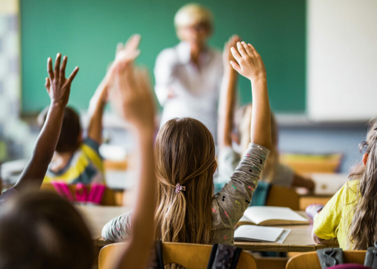 Back view of large group of students raising their hands to answer the question on a class at elementary school.