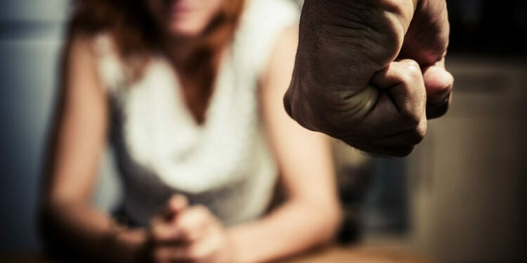 Young woman is sitting hunched at a table at home, the focus is on a man's fist in the foregound of the image